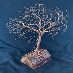 Puede incluir: Una escultura de &aacute;rbol hecha de alambre de cobre fino y retorcido, con ramas intrincadas que se extienden hacia afuera. La base del &aacute;rbol de alambre est&aacute; anclada en una pieza de madera natural semicil&iacute;ndrica con corteza y veta.
