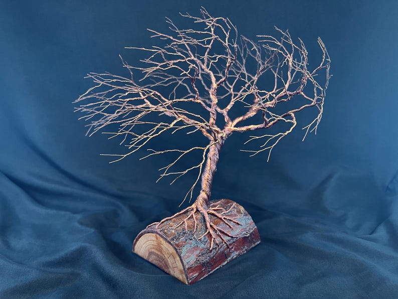 Puede incluir: Una escultura de &aacute;rbol hecha de alambre de cobre fino y retorcido, con ramas intrincadas que se extienden hacia afuera. La base del &aacute;rbol de alambre est&aacute; anclada en una pieza de madera natural semicil&iacute;ndrica con corteza y veta.