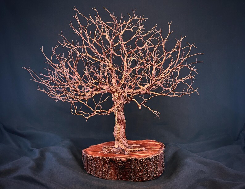 May include: A copper wire sculpture of a leafless tree with intricate, twisted branches and a thick, textured trunk. The tree is mounted on a circular, reddish-brown wooden base with visible bark edges and a flat, polished top surface.
