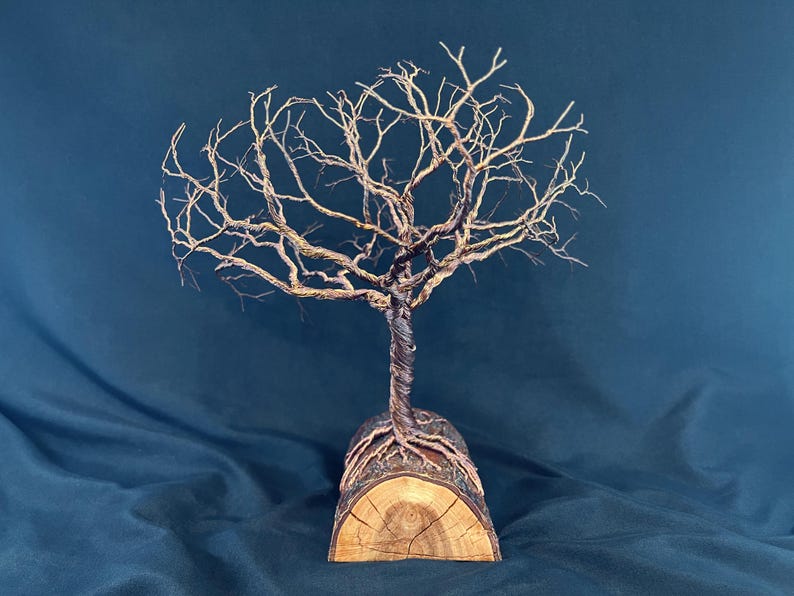 Puede incluir: Una escultura de alambre de un &aacute;rbol sin hojas con ramas met&aacute;licas oscuras retorcidas y un tronco anclado en una base de tronco de madera semicil&iacute;ndrica. El &aacute;rbol presenta finos hilos de alambre que se extienden formando una copa ramificada.