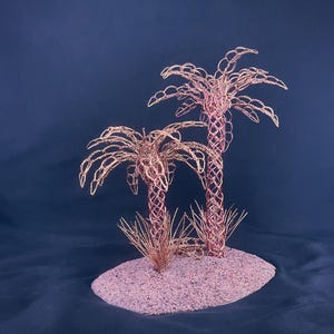 May include: A decorative sculpture featuring two palm trees crafted from thin copper-colored wire. The trees have intricate, woven trunks and arched, leafy fronds, standing on a textured, oval-shaped base covered in fine, metallic copper-toned sand.