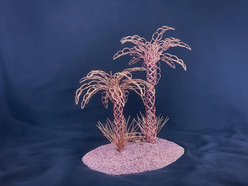 May include: A decorative sculpture featuring two palm trees crafted from thin copper-colored wire. The trees have intricate, woven trunks and arched, leafy fronds, standing on a textured, oval-shaped base covered in fine, metallic copper-toned sand.