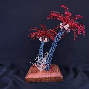 May include: A decorative sculpture featuring two palm trees crafted from blue and red wire, with white spherical accents representing coconuts. The trees are mounted on a square wooden base, accompanied by a small cluster of thin white wire blades.