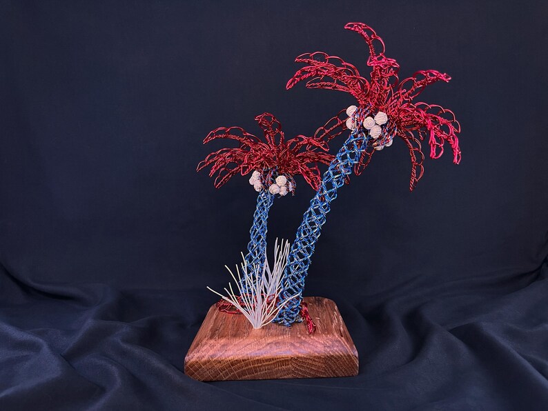 May include: A decorative sculpture featuring two palm trees crafted from blue and red wire, with white spherical accents representing coconuts. The trees are mounted on a square wooden base, accompanied by a small cluster of thin white wire blades.