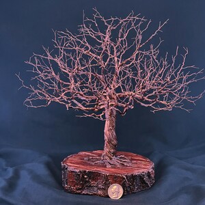May include: A copper wire sculpture of a leafless tree with a twisted trunk and intricate branches, mounted on a circular wooden base. A silver-coloured coin is placed at the base for scale, showing the relative size of the sculpture.
