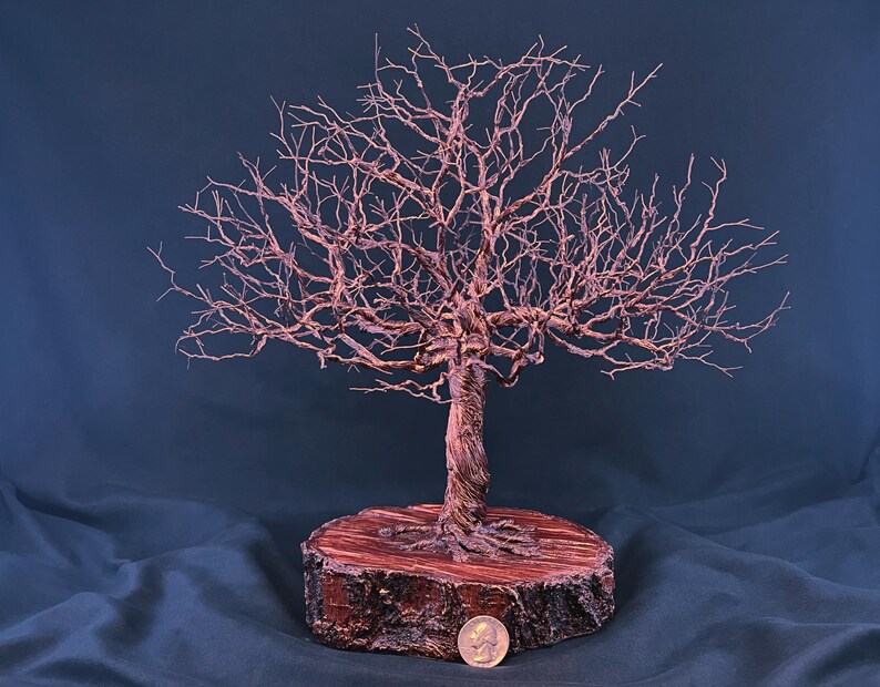 May include: A copper wire sculpture of a leafless tree with a twisted trunk and intricate branches, mounted on a circular wooden base. A silver-coloured coin is placed at the base for scale, showing the relative size of the sculpture.