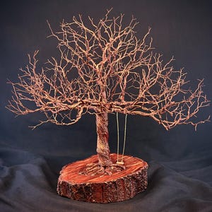 May include: A wire sculpture of a leafless tree with a twisted trunk and intricate branches, mounted on a circular wooden slice base. A small, simple swing hangs from one of the lower branches, featuring two thin vertical cords and a seat.