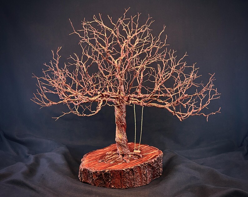 May include: A wire sculpture of a leafless tree with a twisted trunk and intricate branches, mounted on a circular wooden slice base. A small, simple swing hangs from one of the lower branches, featuring two thin vertical cords and a seat.