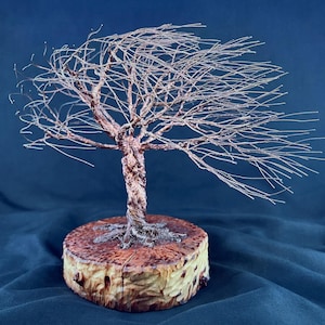May include: A wire sculpture of a tree with thin, metallic branches extending to one side, as if blown by the wind. The trunk is formed from twisted copper-colored wire, and the base is a circular, light-brown wooden slice with visible grain.