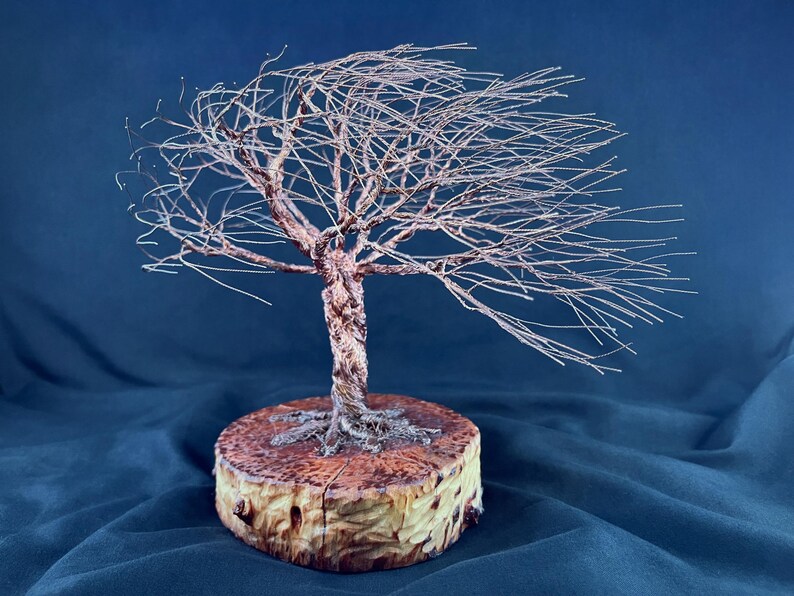 May include: A wire sculpture of a tree with thin, metallic branches extending to one side, as if blown by the wind. The trunk is formed from twisted copper-colored wire, and the base is a circular, light-brown wooden slice with visible grain.