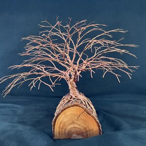 Puede incluir: Una escultura de &aacute;rbol hecha de alambre de cobre fino, con ramas intrincadas que se extienden hacia afuera. La base del &aacute;rbol est&aacute; formada por una pieza de madera natural semicil&iacute;ndrica, que muestra vetas visibles y corteza en el exterior.