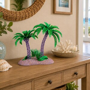 May include: A decorative sculpture featuring two palm trees with green fronds and purple textured trunks, set on a sandy-colored base with small green grass accents. The sculpture is crafted with a detailed, woven-like texture on the trunks.