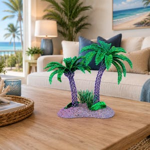 May include: A decorative sculpture featuring two palm trees with vibrant green fronds and textured purple trunks, set upon a small, rounded, light purple base that mimics a sandy island, accented with small green foliage at the base of the trees.