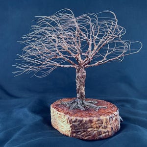 May include: A wire sculpture of a tree with thin, spreading branches and a twisted trunk, anchored to a thick, circular wooden base with visible bark texture. The metal wire has a copper-toned finish, and the tree appears to lean to one side.