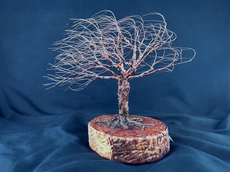 May include: A wire sculpture of a tree with thin, spreading branches and a twisted trunk, anchored to a thick, circular wooden base with visible bark texture. The metal wire has a copper-toned finish, and the tree appears to lean to one side.