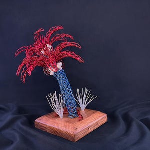 May include: A wire sculpture of a palm tree with a blue woven trunk and red fronds, featuring small white accents at the base of the leaves. The sculpture is mounted on a square wooden base with two small white wire sprigs at the bottom.