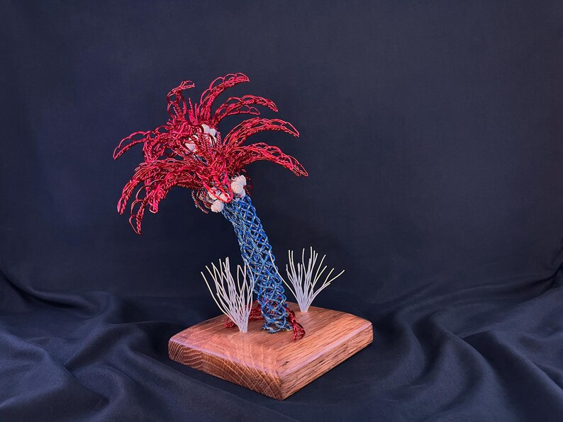 May include: A wire sculpture of a palm tree with a blue woven trunk and red fronds, featuring small white accents at the base of the leaves. The sculpture is mounted on a square wooden base with two small white wire sprigs at the bottom.