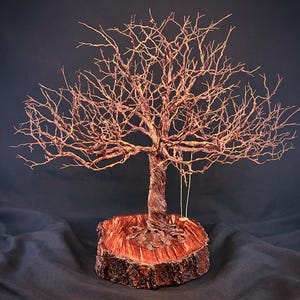 May include: A copper wire sculpture of a leafless tree with intricate branches, mounted on a circular wooden base with a polished reddish-brown surface. A small, simple swing hangs from one of the lower branches of the metallic tree structure.