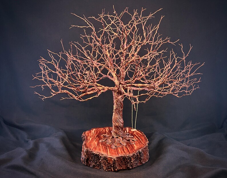 May include: A copper wire sculpture of a leafless tree with intricate branches, mounted on a circular wooden base with a polished reddish-brown surface. A small, simple swing hangs from one of the lower branches of the metallic tree structure.