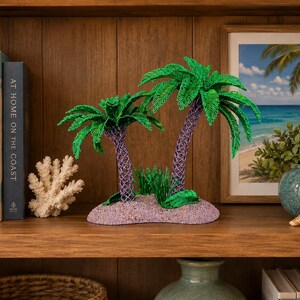 May include: A decorative tabletop sculpture featuring two palm trees with vibrant green fronds and textured purple trunks, set upon a sandy-colored base with small green foliage accents, designed as a miniature tropical island display piece.