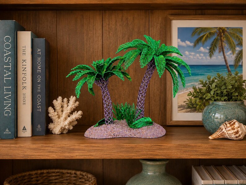 May include: A decorative tabletop sculpture featuring two palm trees with vibrant green fronds and textured purple trunks, set upon a sandy-colored base with small green foliage accents, designed as a miniature tropical island display piece.
