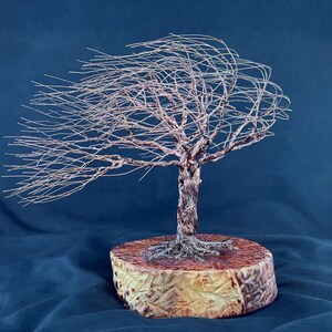 May include: A wire sculpture of a tree with thin, metallic branches extending to one side as if blown by the wind. The tree has a textured, twisted trunk and is mounted on a circular, light-colored wooden base with visible grain patterns.