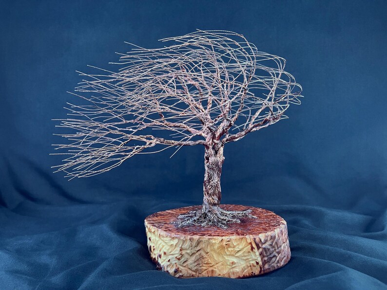 May include: A wire sculpture of a tree with thin, metallic branches extending to one side as if blown by the wind. The tree has a textured, twisted trunk and is mounted on a circular, light-colored wooden base with visible grain patterns.