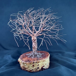 May include: A wire sculpture of a leafless tree with a twisted metallic trunk and thin, spreading branches. The sculpture is mounted on a circular, light-colored wooden base with a natural, textured grain and dark brown markings on the top surface.