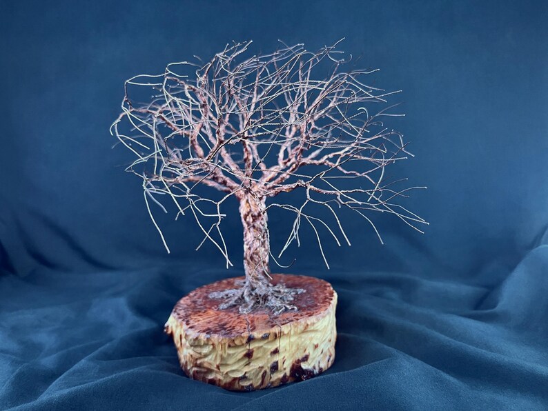 May include: A wire sculpture of a leafless tree with a twisted metallic trunk and thin, spreading branches. The sculpture is mounted on a circular, light-colored wooden base with a natural, textured grain and dark brown markings on the top surface.