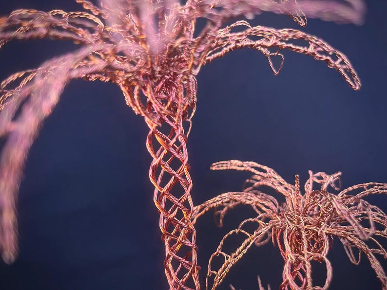 May include: A sculpture of two palm trees crafted from thin, intertwined copper-colored wire. The trunks feature a lattice-like woven pattern, while the fronds are formed by delicate, looping wire strands that extend outward from the top.