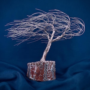 Puede incluir: Una escultura de &aacute;rbol hecha de alambre met&aacute;lico fino de color cobre, con un tronco retorcido y ramas que se extienden hacia un lado como si fueran sopladas por el viento. El &aacute;rbol est&aacute; anclado en una base cil&iacute;ndrica con corteza de color marr&oacute;n rojizo.