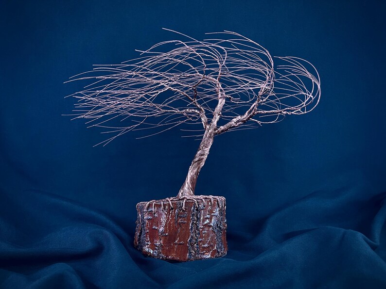 Puede incluir: Una escultura de &aacute;rbol hecha de alambre met&aacute;lico fino de color cobre, con un tronco retorcido y ramas que se extienden hacia un lado como si fueran sopladas por el viento. El &aacute;rbol est&aacute; anclado en una base cil&iacute;ndrica con corteza de color marr&oacute;n rojizo.