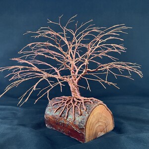 Puede incluir: Una escultura de &aacute;rbol hecha de alambre de cobre fino, con ramas intrincadas y un tronco que se extiende en ra&iacute;ces expuestas. Las ra&iacute;ces est&aacute;n ancladas sobre una pieza cil&iacute;ndrica de madera natural con corteza visible y veta clara.
