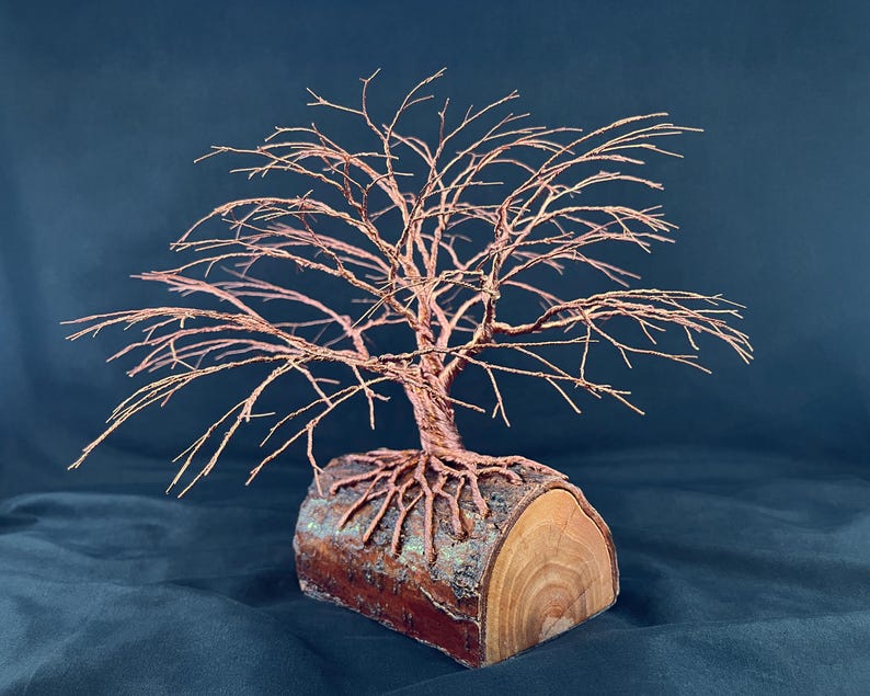 Puede incluir: Una escultura de &aacute;rbol hecha de alambre de cobre fino, con ramas intrincadas y un tronco que se extiende en ra&iacute;ces expuestas. Las ra&iacute;ces est&aacute;n ancladas sobre una pieza cil&iacute;ndrica de madera natural con corteza visible y veta clara.