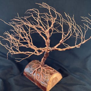 Puede incluir: Una escultura de &aacute;rbol hecha de alambre de cobre trenzado, con ramas intrincadas que se extienden hacia afuera. La base del &aacute;rbol est&aacute; anclada en una pieza cil&iacute;ndrica de madera natural, con ra&iacute;ces de alambre que se extienden por la superficie.