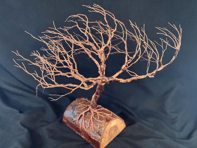 Puede incluir: Una escultura de &aacute;rbol hecha de alambre de cobre trenzado, con ramas intrincadas que se extienden hacia afuera. La base del &aacute;rbol est&aacute; anclada en una pieza cil&iacute;ndrica de madera natural, con ra&iacute;ces de alambre que se extienden por la superficie.