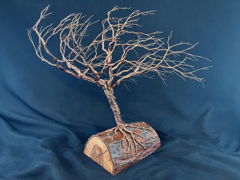 Puede incluir: Una escultura de &aacute;rbol con un tronco met&aacute;lico retorcido y ramas intrincadas y extendidas. El &aacute;rbol est&aacute; anclado a una pieza de madera natural semicil&iacute;ndrica que sirve de base, con ra&iacute;ces met&aacute;licas que se extienden sobre la superficie.