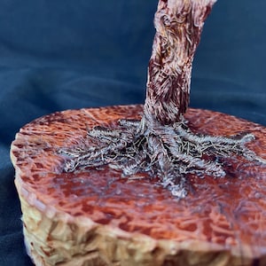 May include: A sculpture of a tree trunk and exposed roots crafted from twisted copper-colored wire. The base is a circular, polished wooden slice with a reddish-brown, glossy finish that highlights the natural grain and texture of the wood.