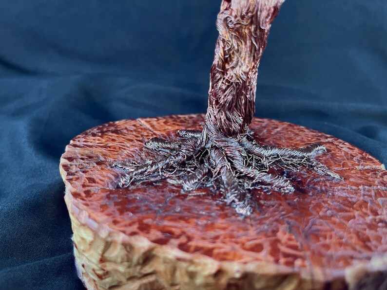 May include: A sculpture of a tree trunk and exposed roots crafted from twisted copper-colored wire. The base is a circular, polished wooden slice with a reddish-brown, glossy finish that highlights the natural grain and texture of the wood.