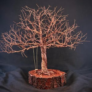 May include: A wire sculpture of a leafless tree with a twisted trunk and intricate branches, featuring a small swing hanging from one branch. The sculpture is mounted on a circular wooden base with visible bark texture on the outer edge.