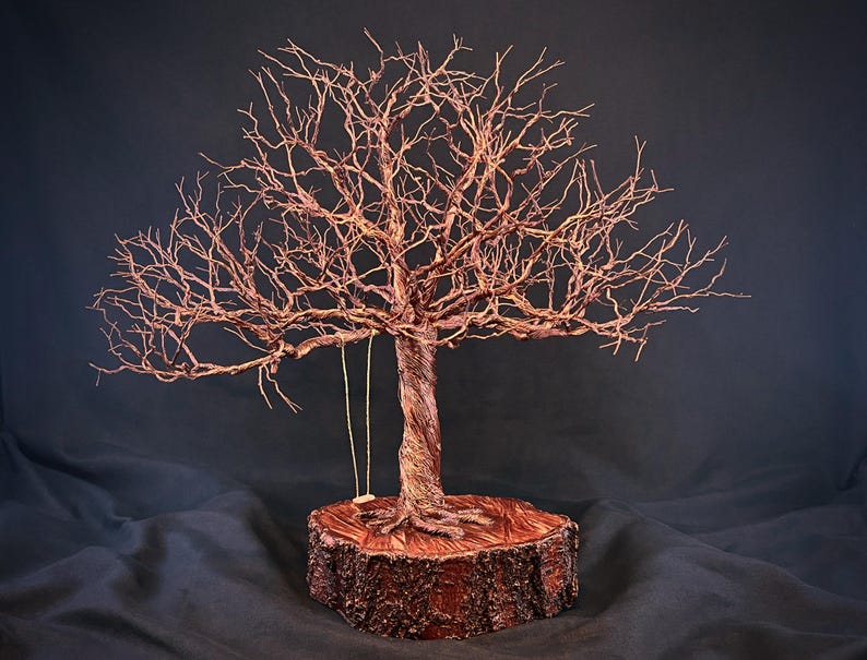 May include: A wire sculpture of a leafless tree with a twisted trunk and intricate branches, featuring a small swing hanging from one branch. The sculpture is mounted on a circular wooden base with visible bark texture on the outer edge.