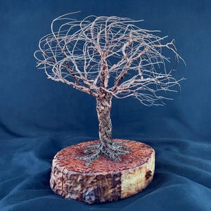 May include: A wire sculpture of a tree with a twisted trunk and thin, spreading branches. The sculpture is mounted on a circular, light brown wooden base with a natural bark edge, showing a textured grain and a warm, polished finish.