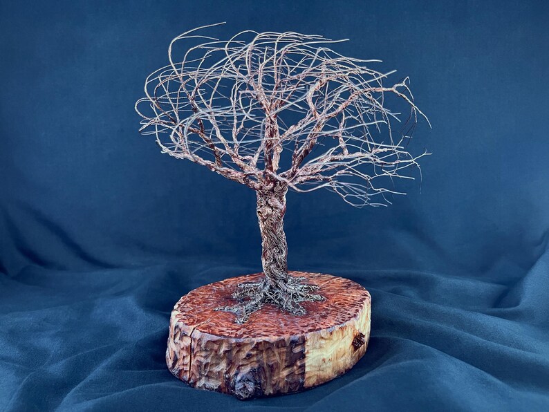 May include: A wire sculpture of a tree with a twisted trunk and thin, spreading branches. The sculpture is mounted on a circular, light brown wooden base with a natural bark edge, showing a textured grain and a warm, polished finish.