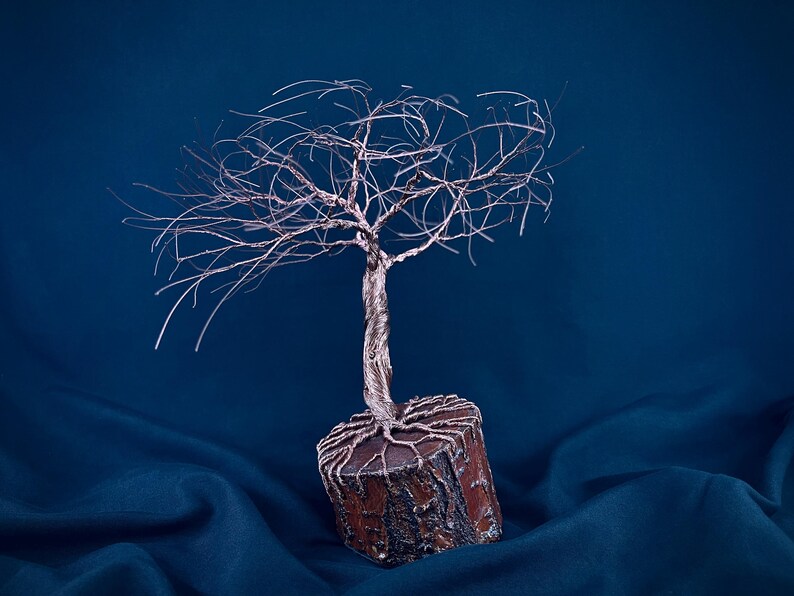 Puede incluir: Una escultura decorativa de &aacute;rbol hecha de alambre fino y retorcido de color cobre. Las ramas de alambre se extienden hacia afuera desde un tronco central anclado en una base cil&iacute;ndrica de madera con textura de corteza y ra&iacute;ces visibles.