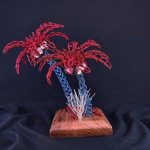 May include: A sculpture of two palm trees crafted from red and blue wire, featuring small white stones nestled in the foliage. The trees are mounted on a square wooden base, with a small cluster of white wire sprigs at the base of the trunks.