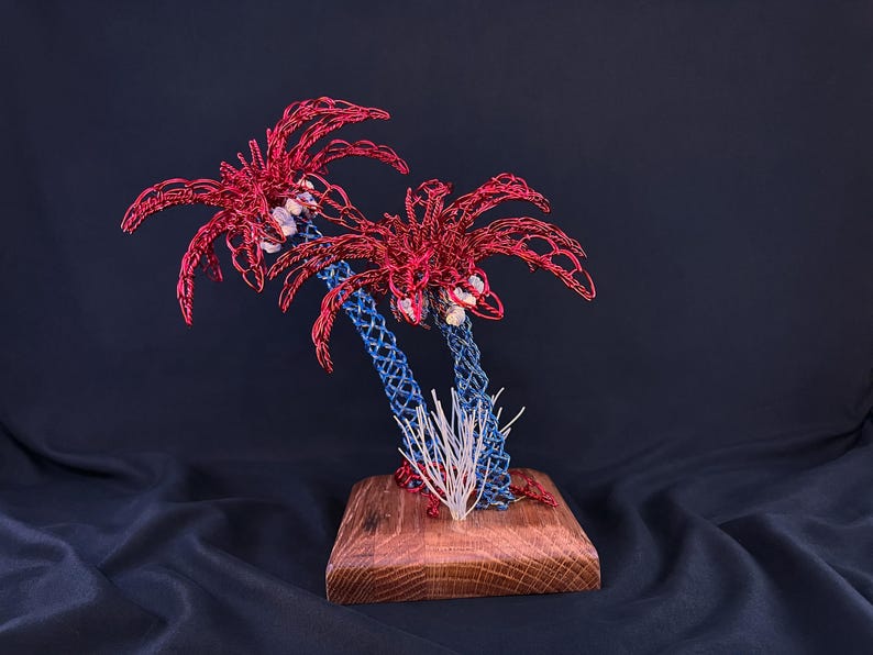 May include: A sculpture of two palm trees crafted from red and blue wire, featuring small white stones nestled in the foliage. The trees are mounted on a square wooden base, with a small cluster of white wire sprigs at the base of the trunks.