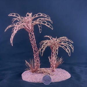 May include: Two palm tree sculptures crafted from thin copper wire, featuring intricate woven trunks and delicate fronds. The trees are mounted on a circular base covered in light-colored sand, with a silver US quarter coin placed for scale.