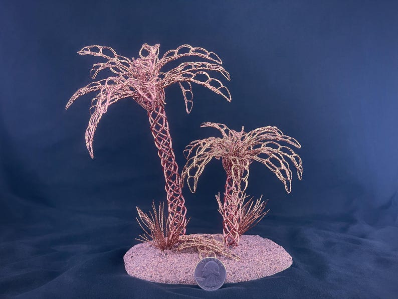 May include: Two palm tree sculptures crafted from thin copper wire, featuring intricate woven trunks and delicate fronds. The trees are mounted on a circular base covered in light-colored sand, with a silver US quarter coin placed for scale.