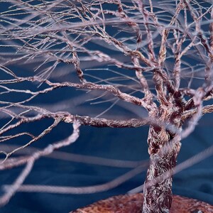 May include: A sculpture of a tree crafted from twisted copper wire, featuring a thick, braided trunk that branches out into numerous thin, delicate limbs. The metallic copper material has a warm, reddish-brown tone set against a dark blue background.