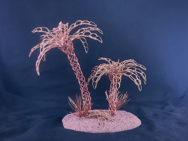 May include: Two palm tree sculptures crafted from thin copper wire, featuring intricate lattice-patterned trunks and arched fronds. The trees are mounted on a circular, textured base with small wire tufts at the base of each trunk.
